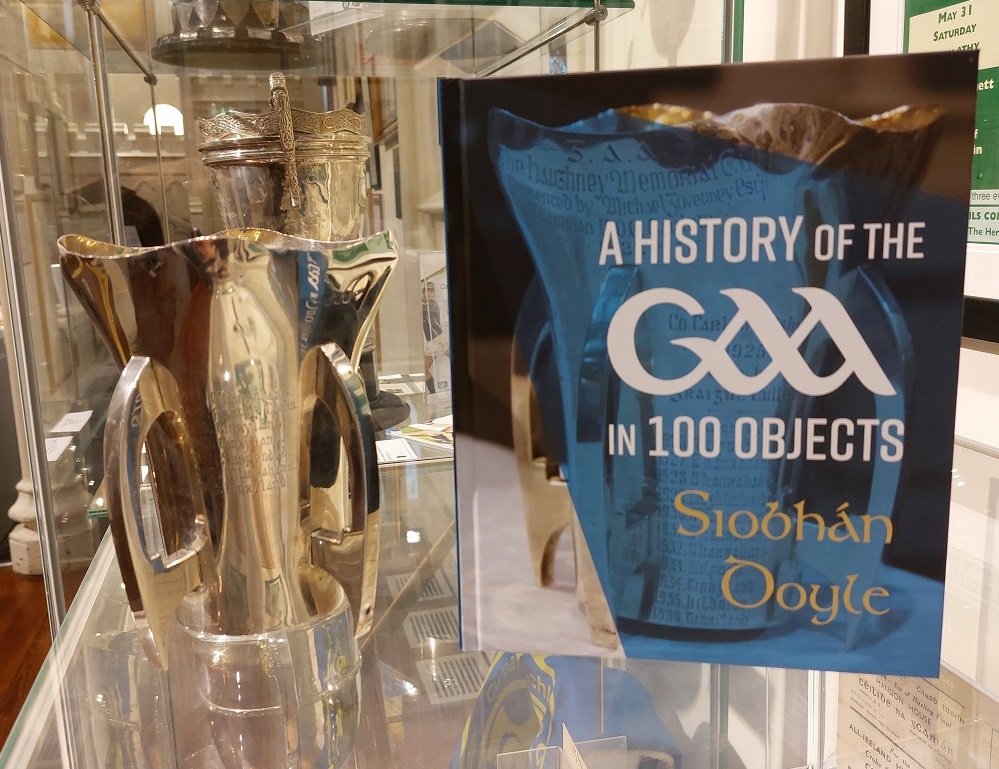 The Haughney GAA Cup, Carlow, beside the front cover of the book A History of the GAA in 100 Objects which features the Haughney Cup on the front cover